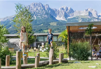 Two children are playing on an obstacle course made of wooden logs in a green garden. In the background, majestic mountains can be seen under a blue sky.