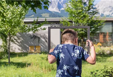 A boy wearing a top with a palm motif is holding a picture frame. In the background, trees and mountains can be seen.