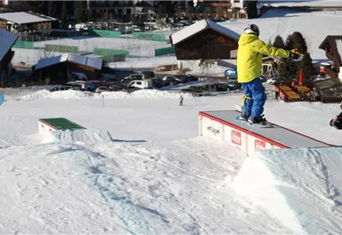 Ein Snowboarder fährt über ein Rails im Schnee. Im Hintergrund sind verschneite Hütten und eine Skipiste zu sehen.