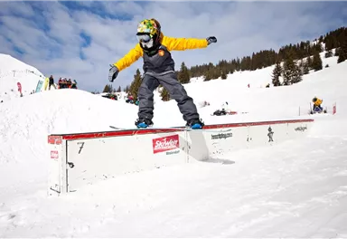 Ein junger Skifahrer führt Tricks auf einem Schneehang vor. Um ihn herum sind andere Sportler und eine verschneite Landschaft zu sehen.