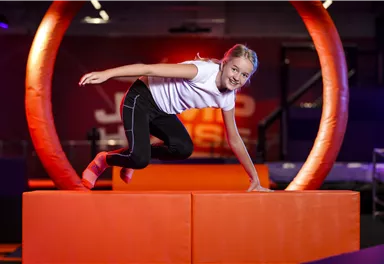 A girl jumps over an orange platform in a trampoline park. The background is colorfully and invitingly designed.