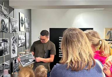 Ein Mann erklärt die Ausstellung mit Fotografien an der Wand. Um ihn herum stehen interessierte Besucher.