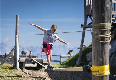 Ein Mädchen balanciert auf einem Seil in den Bergen. Im Hintergrund sind eine schöne Landschaft und eine Holzstruktur sichtbar.