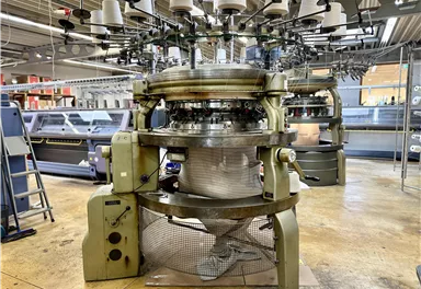Eine Strickmaschine in einer Fabrik. Sie ist groß und hat mehrere Spulen oben, die zum Stricken verwendet werden.