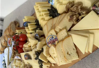 Eine Auswahl an verschiedenen Käsesorten auf einem Holzbrett. Dazu liegen Tomaten, Nüsse und Brot.