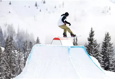 Ein Snowboarder springt über ein gestyltes Snowpark-Element in einer verschneiten Landschaft. Im Hintergrund sind Tannenbäume und Berggipfel sichtbar.