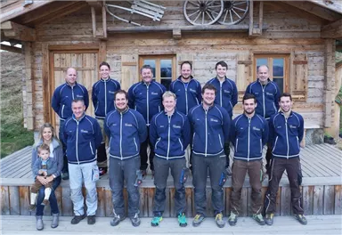 Eine Gruppe von Männern in blauen Jacken steht vor einer Holzhütte. Eine Frau mit einem Kind sitzt in der unteren Reihe.