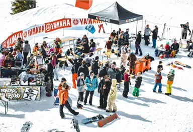 Eine belebte Winterveranstaltung mit vielen Menschen im Schnee. Tische und Stände sind aufgestellt, und die Teilnehmer genießen die Atmosphäre.