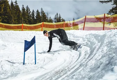 Ein Snowboarder fährt durch den Schnee und hat eine Kurve genommen. Im Hintergrund sind schneebedeckte Bäume und eine gelbe Absperrung zu sehen.