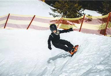 Ein Junge fährt auf einem Snowboard im Schnee. Im Hintergrund sind Sicherheitsnetze zu sehen.