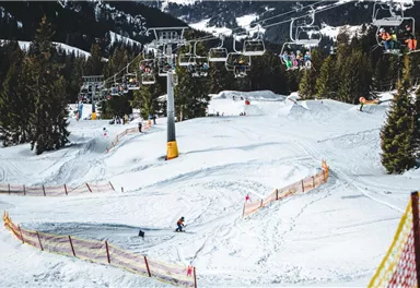 Eine verschneite Berglandschaft mit Skifahrern auf der Piste. Im Hintergrund sind Sessellifte und hohe Tannenbäume zu sehen.