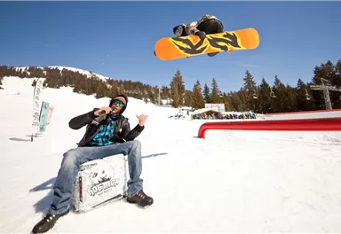 Ein Snowboarder springt hoch in die Luft über einem Schneehang. Im Vordergrund sitzt ein Mann und beobachtet die Aktion.