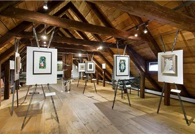 An expanded attic with wooden ceilings. Several artworks in frames are displayed on stands.