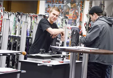 A ski shop with two salespeople discussing ski equipment. In the background, many skis are visible.