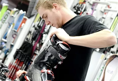 A man is fitting a ski boot in a sports store. In the background, skis and other equipment are visible.