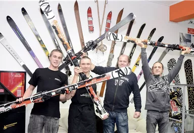 Four men are standing in the shop, surrounded by skis on the wall. They are holding different skis in their hands and appear excited.