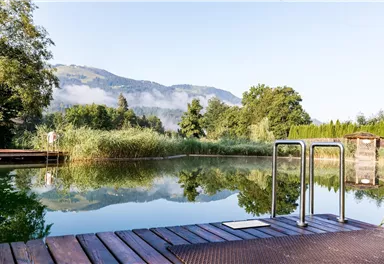 A tranquil natural lake with green shores and gentle mountains in the background. The surface of the water reflects the surrounding landscape.