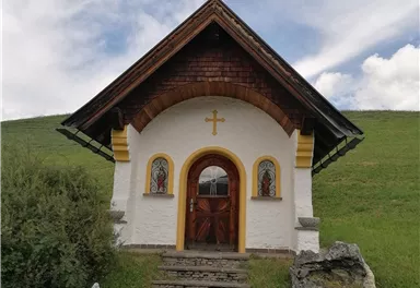 A small, charming chapel with a wooden roof and colorful windows stands on a green meadow. The sky is slightly cloudy.