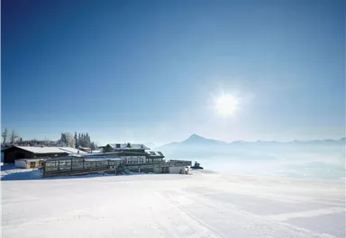A picturesque winter landscape with snow-covered ground and clear blue sky. In the background, mountains can be seen while the sun is shining.