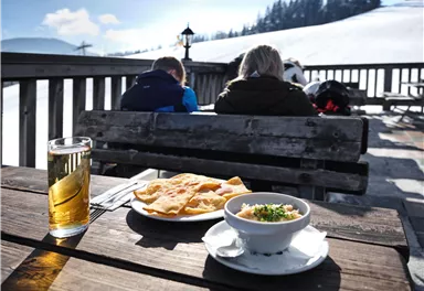 A table outdoors with a view of snow-covered mountains. On it sits a glass of beer, a plate with flatbread, and a bowl with a warm dish.