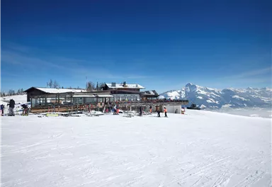A mountainous winter landscape with plenty of snow and a clear blue sky. In the foreground stands a ski lodge, surrounded by skiers.