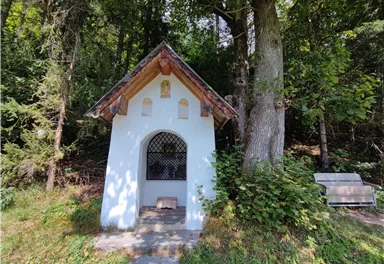 A small chapel stands in a wooded area. Next to the chapel is a wooden bench.