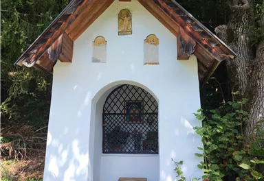 A small chapel in nature, surrounded by trees. The white wall and the simple roof give it a charming appearance.