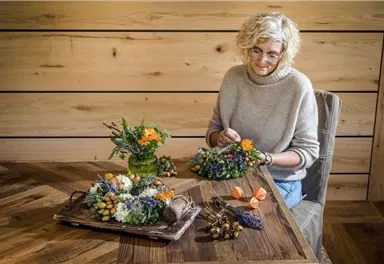 Eine Frau arbeitet an einem Tisch mit verschiedenen Blumenarrangements. Im Hintergrund sind Holzflächen sichtbar, was eine gemütliche Atmosphäre schafft.