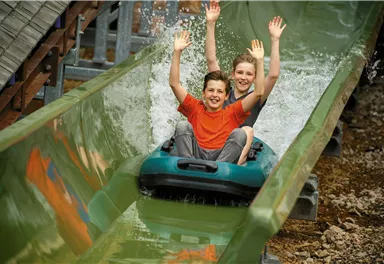 Two children are having a lot of fun on a water slide. They are sitting in an inflatable ring and splashing water as they slide down.