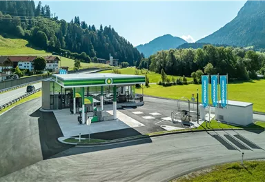 A modern gas station in a picturesque mountain landscape. The surroundings are green with hills and blue mountains in the background.