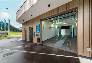 A modern car wash with a large entrance and a bright, clean interior view. In the background, green meadows and mountains can be seen.