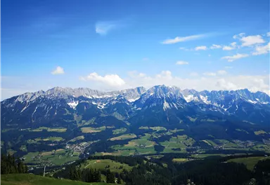 An impressive mountain landscape with snow-capped peaks and green meadows. The sky is clear with a few clouds.