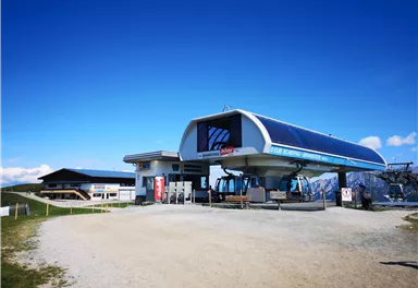 A modern cable car station in a mountainous landscape. The sky is clear and blue, surrounded by green meadows.