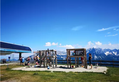A playground in the mountains with a view of snow-capped peaks. Several children play on the wooden play structure under a bright blue sky.