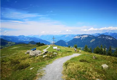 A view of the mountains with a hiking trail and a cross in the distance. The sky is clear and a green, wooded landscape can be seen.