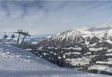 A snow-covered mountain landscape with a ski lift and clear, blue skies. In the background, majestic mountains and valleys can be seen.