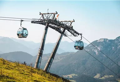 A cable car with gondolas on a mountain. In the background, impressive, forested mountains can be seen.