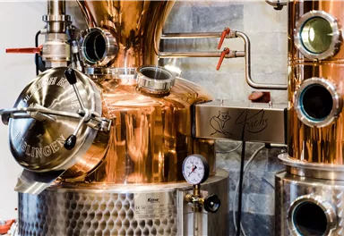 A copper distillation apparatus with various pipes and a pressure gauge. In the background, stone walls are visible.