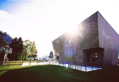 Ein modernes Gebäude mit Holzverkleidung und schrägem Dach. Im Hintergrund sind Bäume und eine Seilbahn zu sehen, während die Sonne hell scheint.