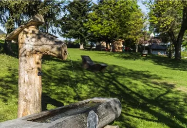A rustic wooden fountain with flowing water stands in a green park. In the background, there are trees and a peaceful meadow.