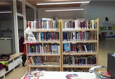 A bookshelf with many different books stands in a room. In the background, tables and a wall with colorful pictures are visible.