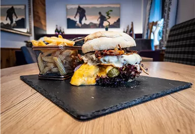 Ein saftiger Burger mit Käse und frischem Gemüse liegt auf einem schwarzen Servierteller. Dazu gibt es knusprige Pommes frites in einem kleinen Korb.