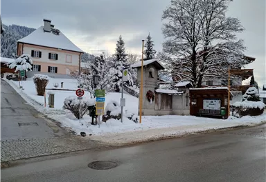 Eine verschneite Straße in einem ruhigen Dorf. Im Hintergrund sind Häuser und verschneite Bäume zu sehen.