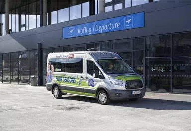 Ein Linienbus steht vor einem modernen Flughafengebäude. Über dem Bus ist das Schild "Abflug / Departure" sichtbar.