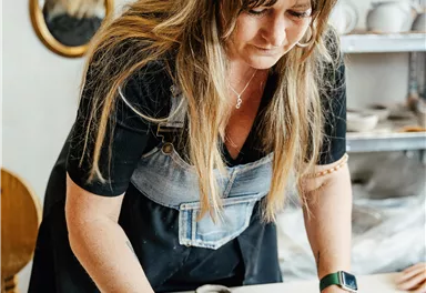 A woman is working focused at the table with clay. She is creatively shaping artworks in a bright workshop.