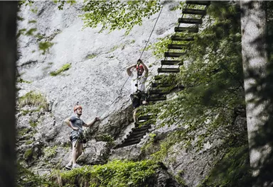 Zwei Personen beim Klettern an einer Felswand mit einer Holzleiter. Umgeben von Bäumen und natürlicher Vegetation.