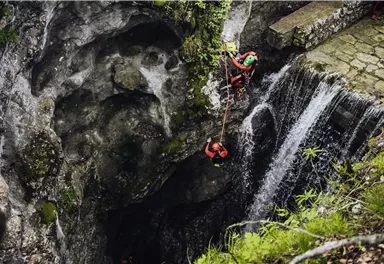 Zwei Personen in Schutzkleidung klettern an einer Felswand. Im Hintergrund fließt ein Wasserfall in eine Schlucht.