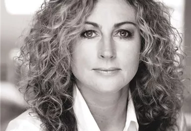 A woman with curly hair is looking directly at the camera. She is wearing a white shirt and has a neutral expression on her face.