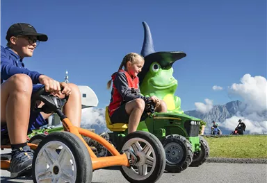 Zwei Kinder fahren mit kleinen Fahrzeugen auf einer sonnigen Wiese. Im Hintergrund ist eine große grüne Figur mit einem Zauberhut zu sehen.