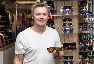 Ein Mann hält eine modische Sonnenbrille in der Hand. Im Hintergrund sind viele weitere Sonnenbrillen in einem Verkaufsregal zu sehen.